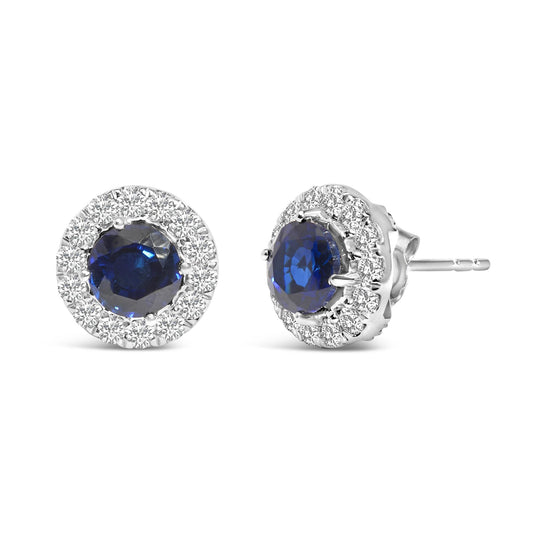 Close-up image of a pair of white stud earrings with round blue gemstones in the center, surrounded by multiple small white gemstones in a prong setting, shown from front and side angles on a white background.