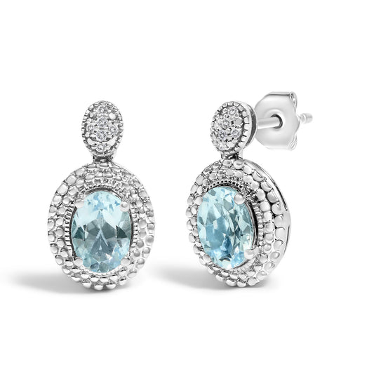 Pair of white metal oval drop earrings with blue oval gemstones surrounded by small white round gemstones in pave setting, shown from a front and side angle on a white background.