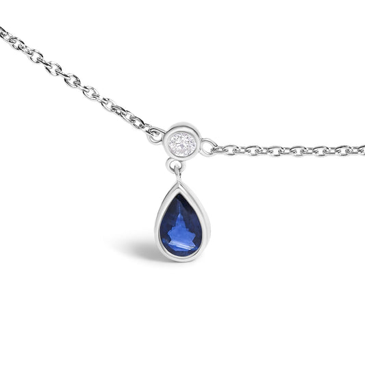 Close-up front view of a white metal necklace chain with a round white gemstone set in bezel, from which a pear-shaped blue gemstone in bezel setting hangs as a pendant.