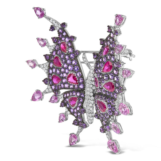 Close-up front angle of a white metal butterfly-shaped brooch adorned with red pear-shaped gemstones, purple round gemstones, pink pear-shaped gemstones, and white round gemstones in various settings.
