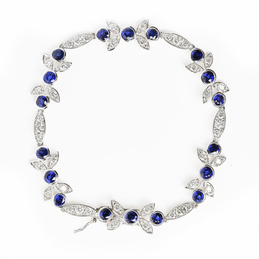 Top-down view of a white metal bracelet with round blue gemstones in bezel settings and round white gemstones in prong settings arranged in leaf and marquise shapes.