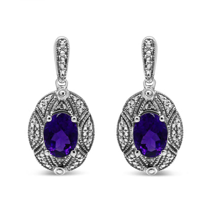 Front view of white metal drop earrings with oval purple gemstones surrounded by intricate detailing with small round white gemstones in prong settings.