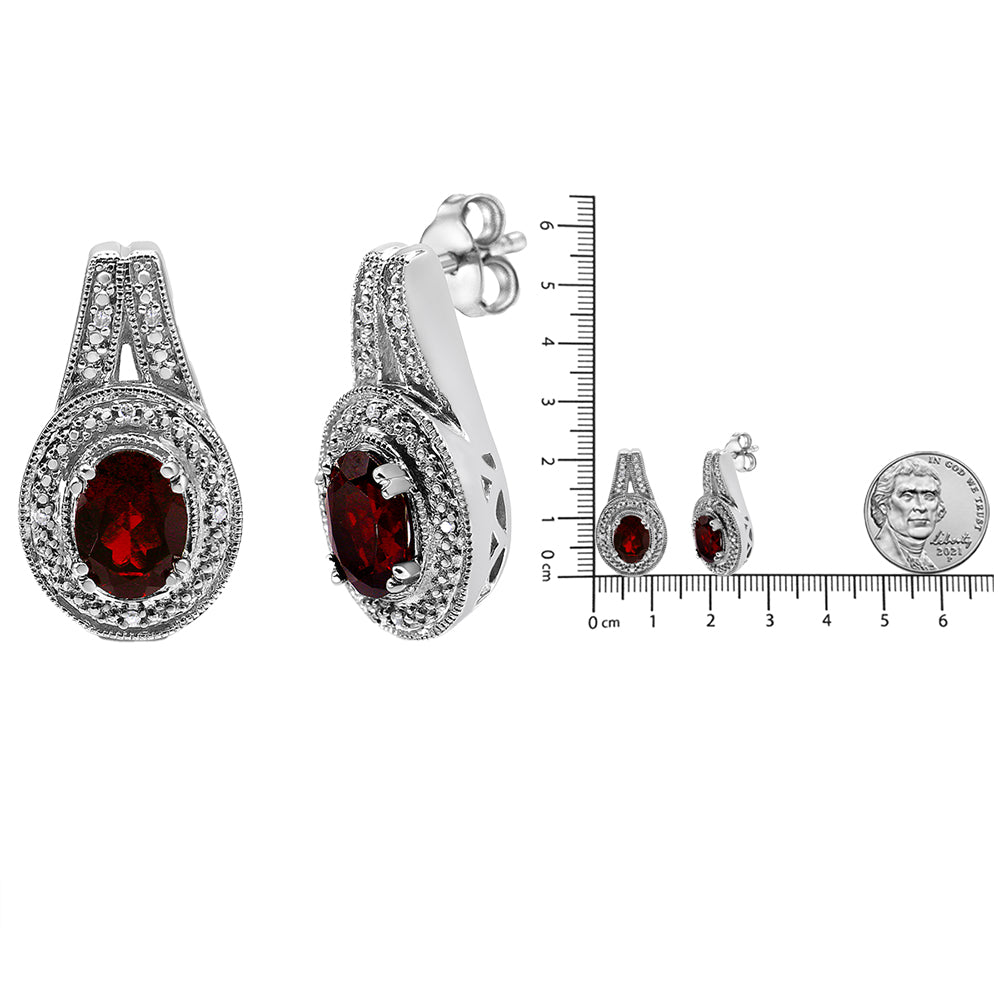 Pair of white metal earrings with oval red gemstones surrounded by round white gemstones, shown front and side views next to a ruler and coin for size reference.