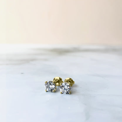 Pair of yellow metal stud earrings with round white gemstones in four-prong settings, shown from a front low angle on a marble surface.