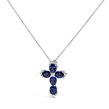 White metal necklace with a cross-shaped pendant featuring oval blue gemstones and round white gemstones in shared prong settings, shown in a front-facing close-up shot.