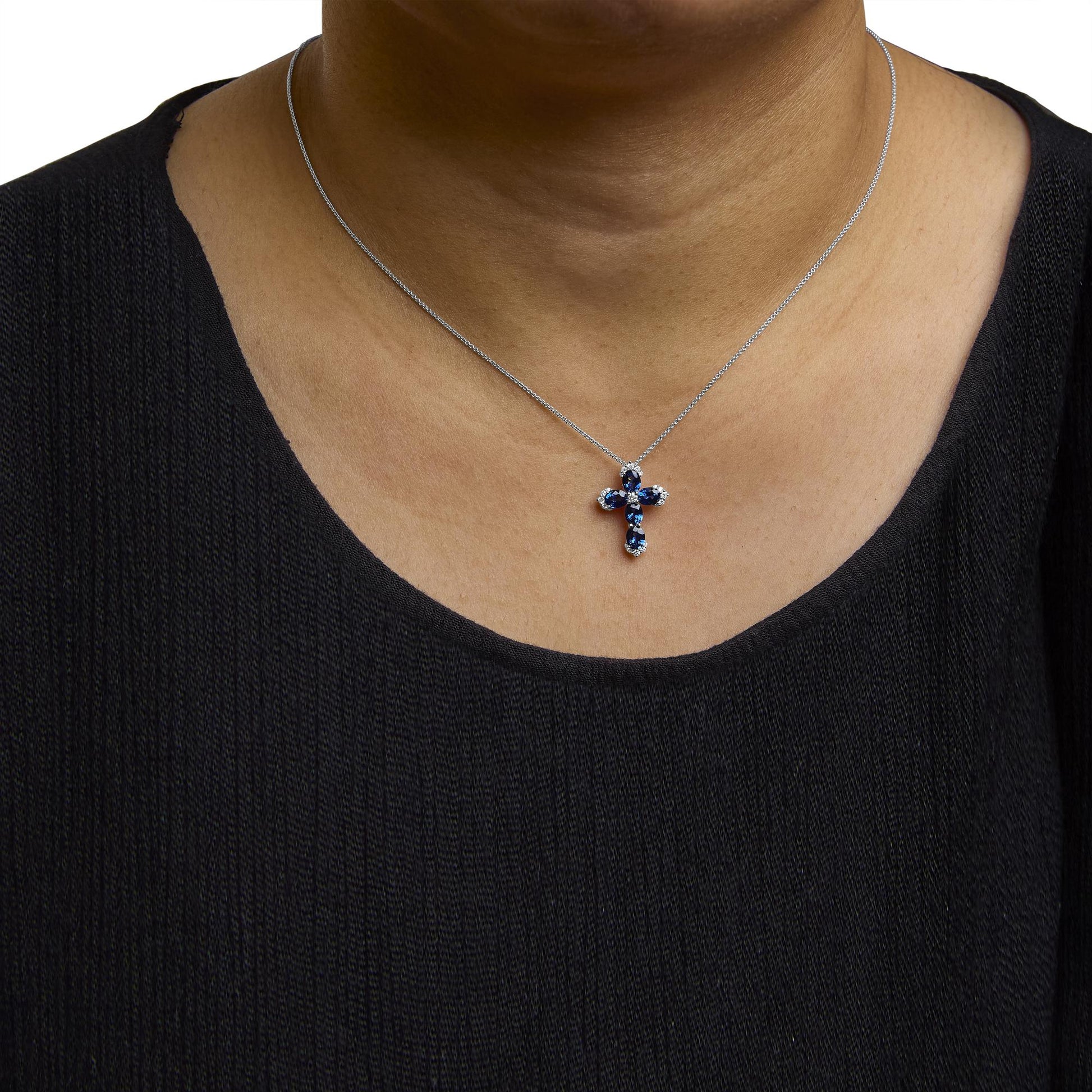 Necklace with a cross-shaped pendant featuring oval blue gemstones and round white gemstones, set in white metal, shown worn on a model from the front.