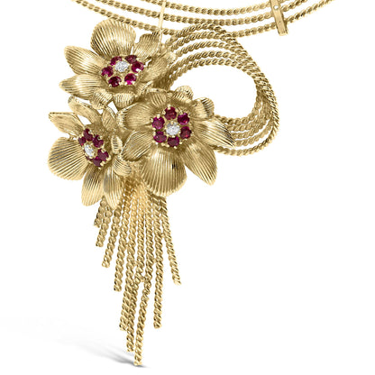 Close-up front angle of a yellow metal necklace featuring three textured flower designs with red gemstones and white gemstones in the center, along with twisted chain tassels hanging below.