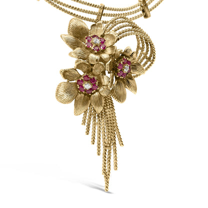 Close-up front angle of a yellow metal necklace with three textured flower pendants, each featuring a cluster of red and white round gemstones in the center, and multiple twisted chain tassels hanging below.