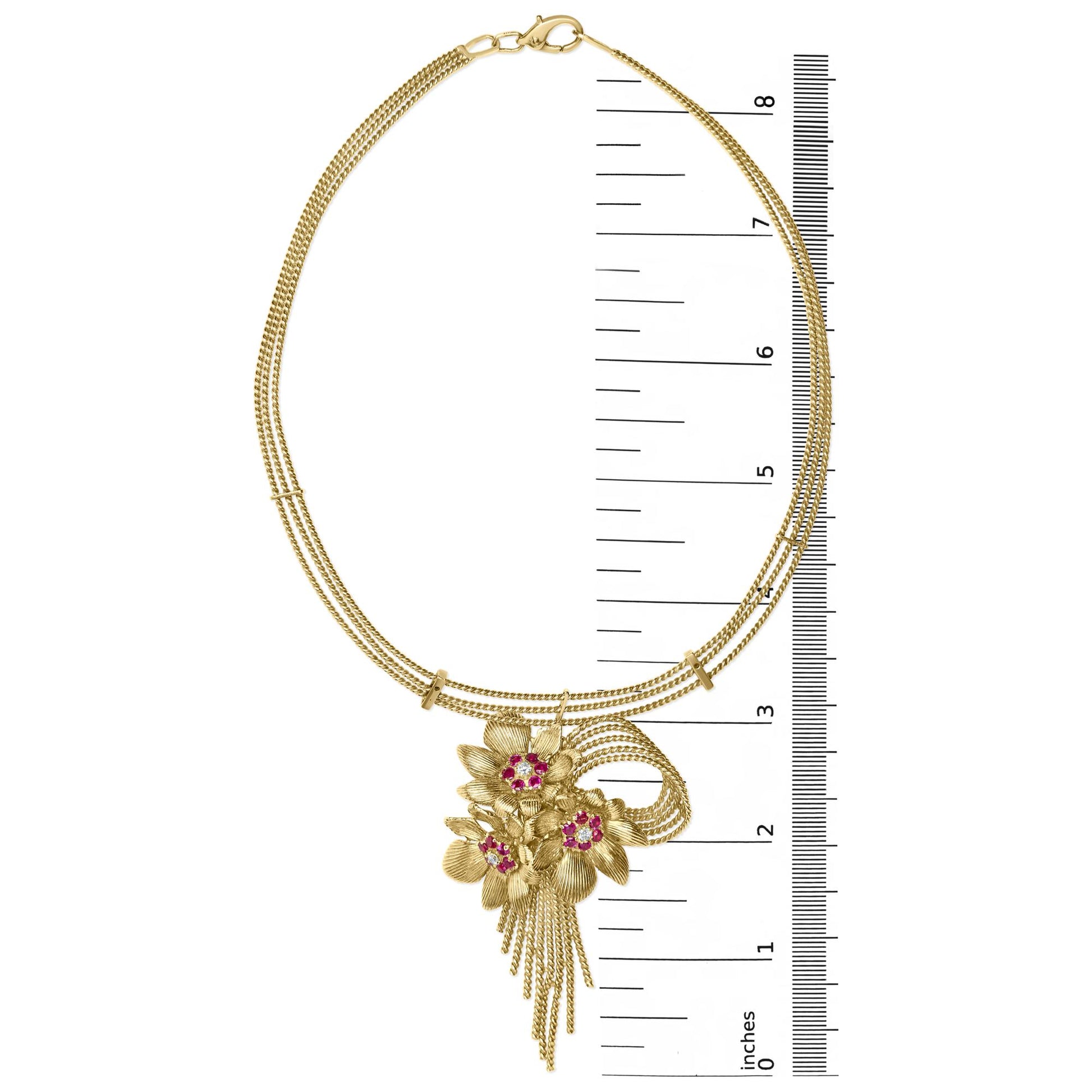 Top-down view of a yellow metal necklace with a floral pendant set with white and red gemstones, shown on a ruler for size reference.