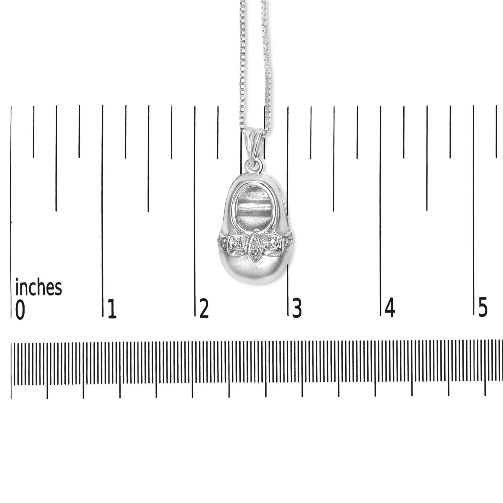 White metal necklace with a pendant shaped like a baby shoe, featuring small round white gemstones in a 4-prong setting across the front, shown hanging on a scale against a white background, front view.