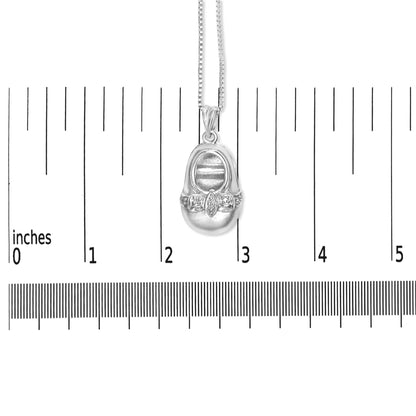 White metal necklace with a pendant shaped like a baby shoe, featuring small round white gemstones in a 4-prong setting across the front, shown hanging on a scale against a white background, front view.