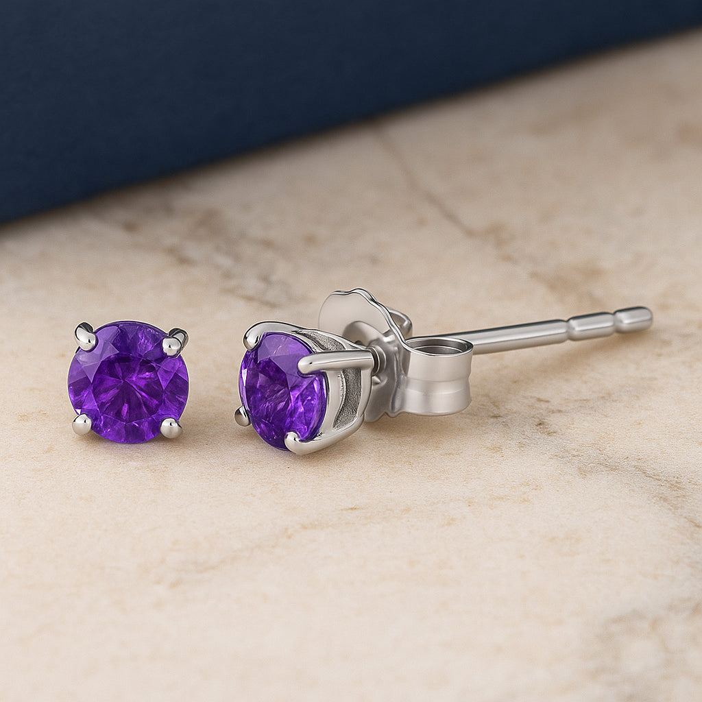 Pair of white metal stud earrings with round purple gemstones in four-prong settings, shown side view on a flat surface with a slight angle.