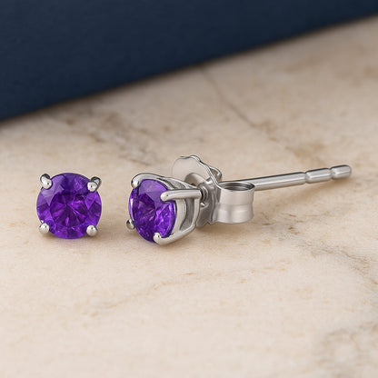 Pair of white metal stud earrings with round purple gemstones in four-prong settings, shown side view on a flat surface with a slight angle.