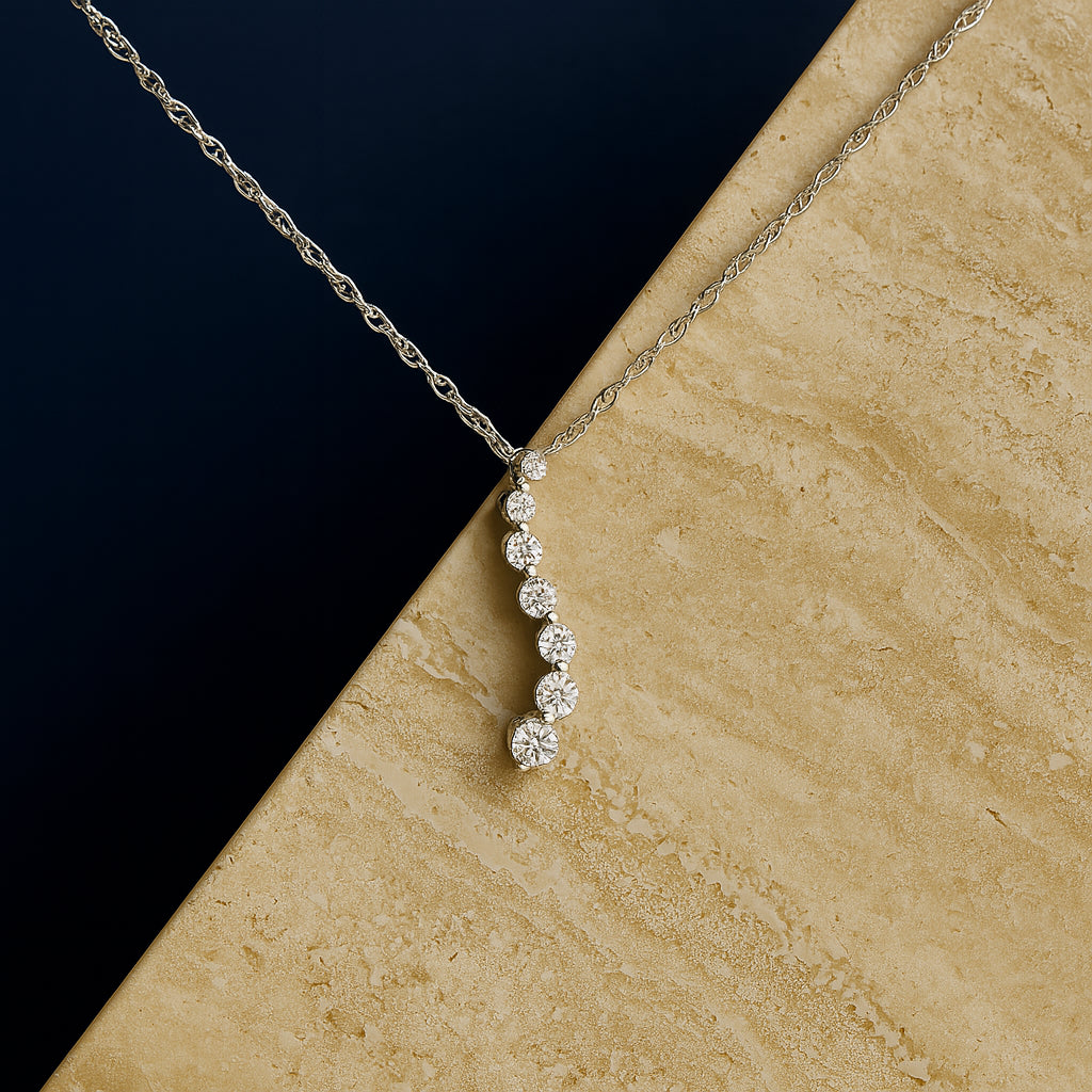 White metal necklace shown against a tan and dark background, featuring a vertical pendant with seven round white gemstones in a prong setting, shot from a slightly angled top view.