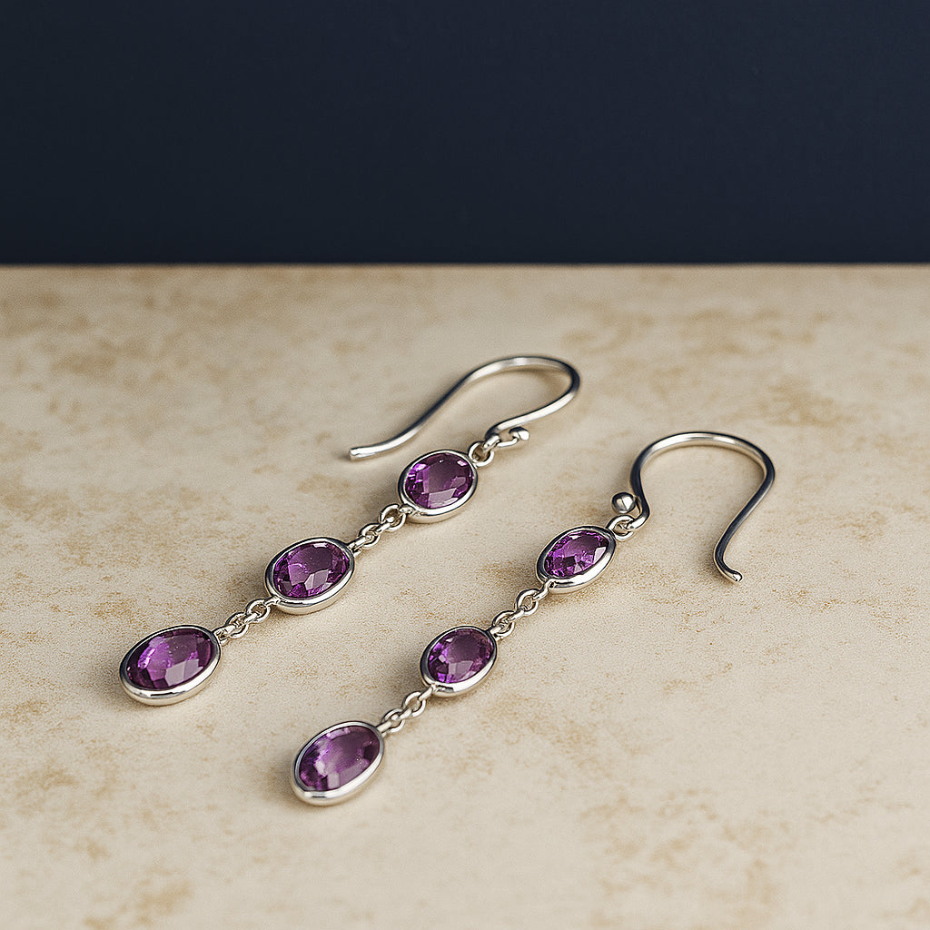Pair of white metal dangle earrings featuring three bezel-set oval purple gemstones in a vertical arrangement, shown in a close-up shot from above.