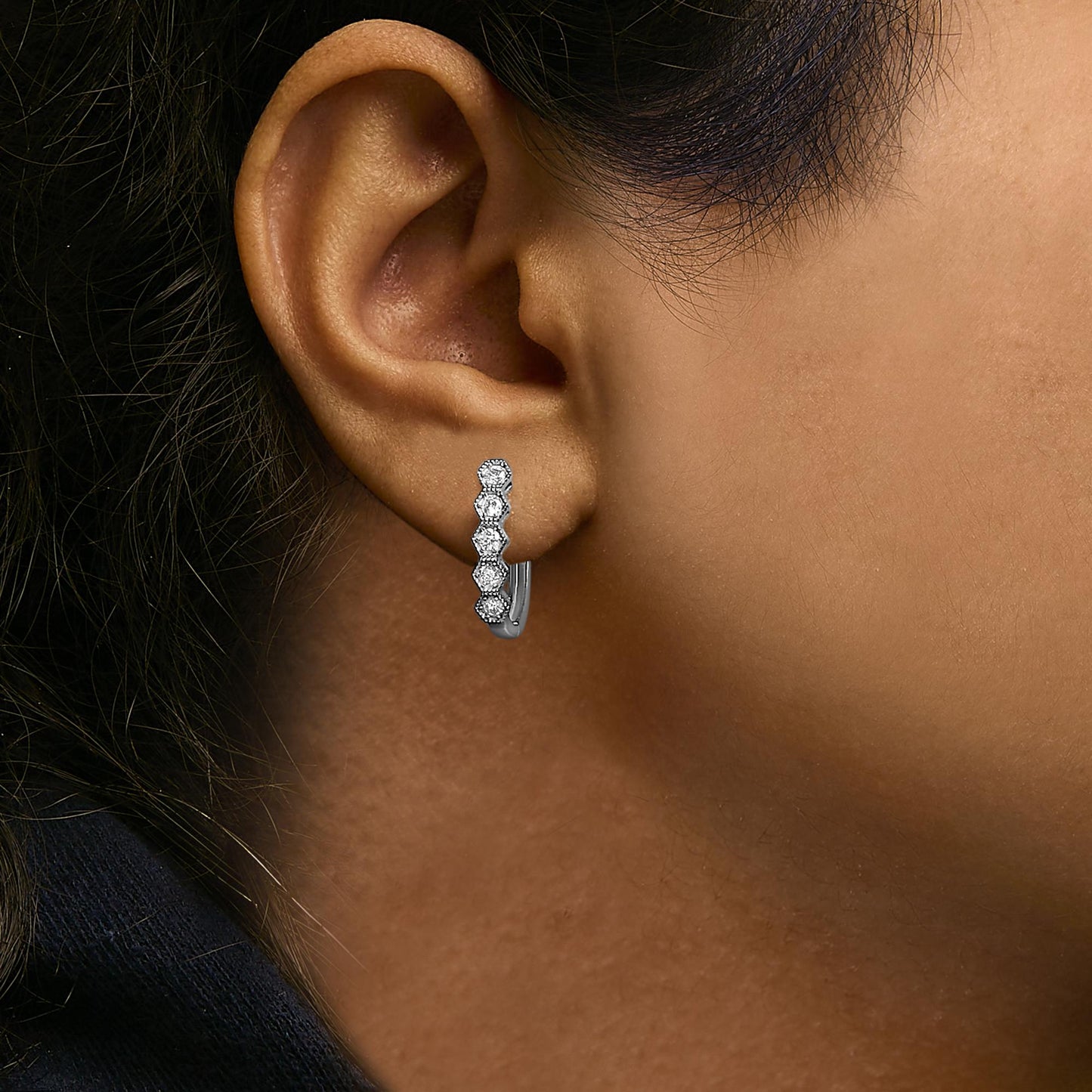 Close-up side view of a white metal hoop earring worn on a model's ear, featuring a row of round white gemstones in six-prong settings along the front.