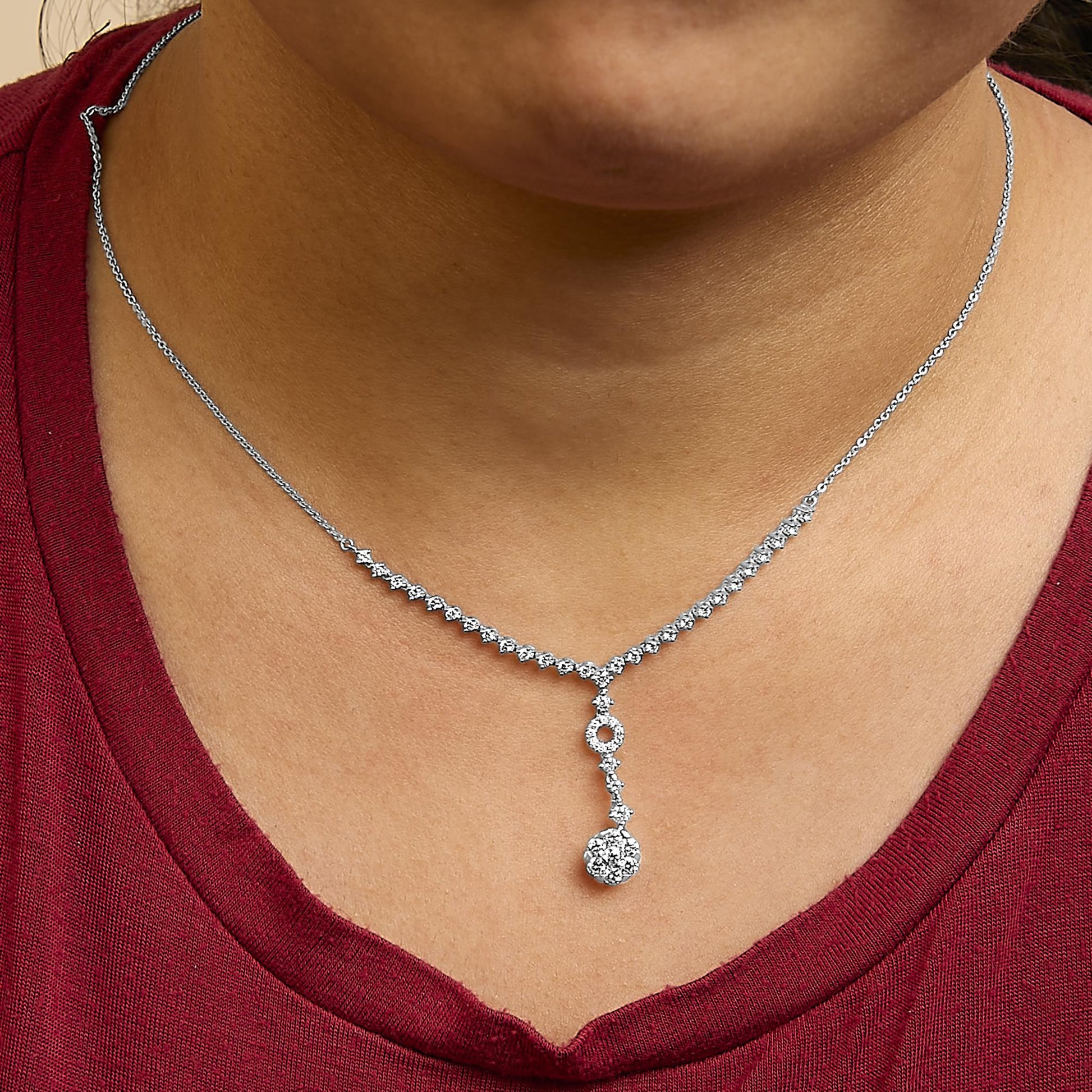White metal necklace shown worn on a model, featuring a chain with a line of small round white gemstones leading to a vertical drop pendant with additional round white gemstones and a cluster of round white gemstones at the end, photographed from the front.