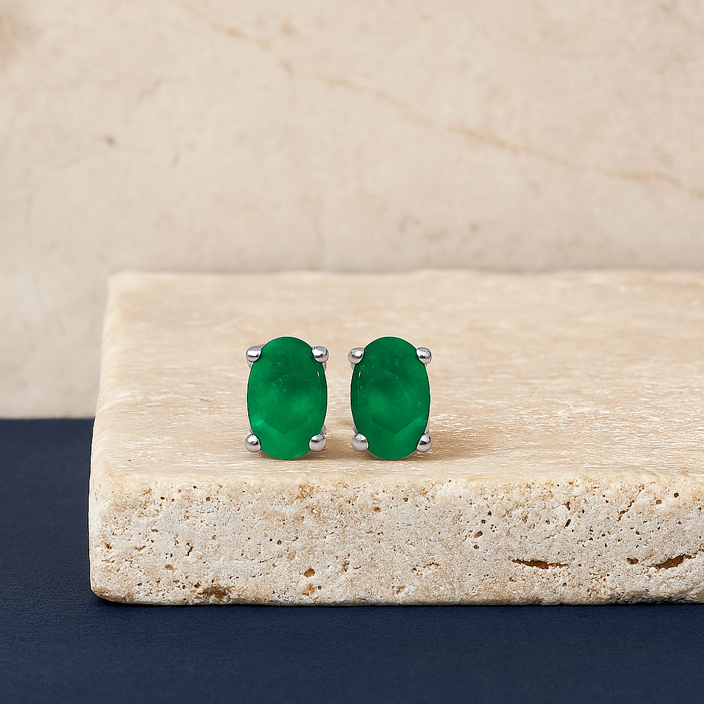 White metal oval green gemstone stud earrings shown on a beige stone surface from the front angle.