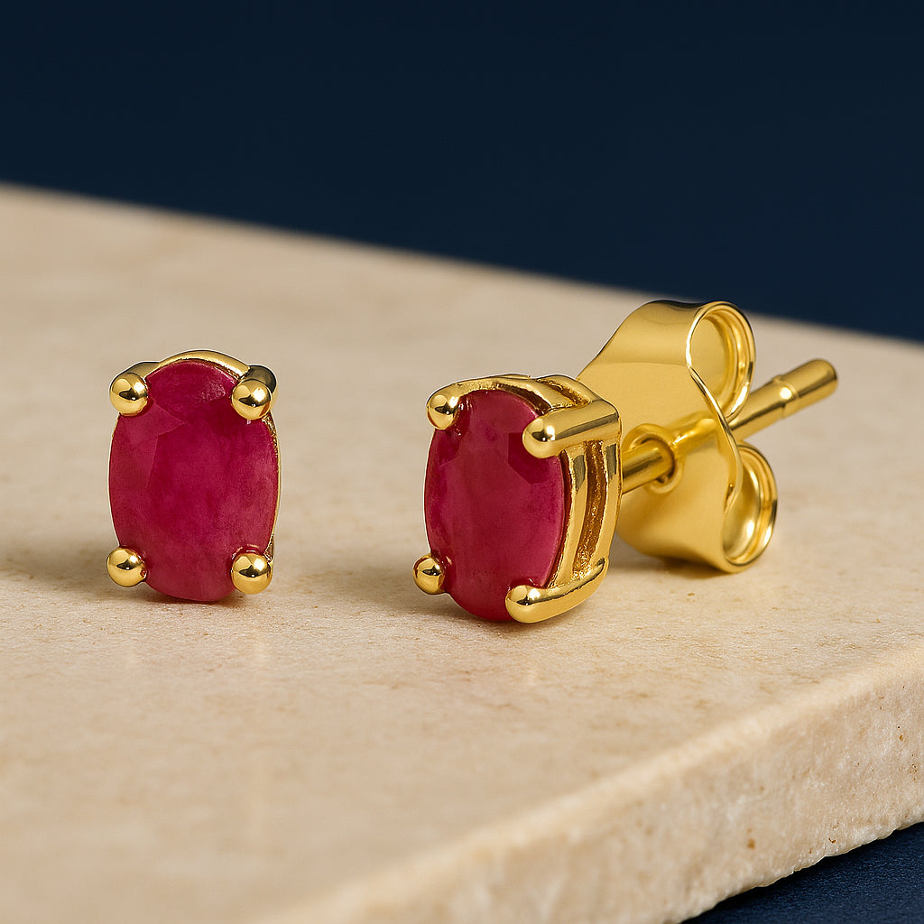 Close-up front and side angle of yellow metal stud earrings with oval-shaped red gemstones in prong settings, displayed on a beige surface.