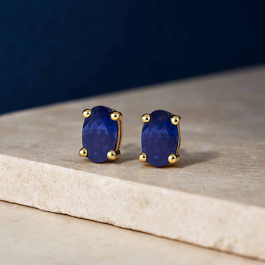 Pair of yellow metal stud earrings with oval-shaped blue gemstones in prong settings, shown from a front angle on a textured beige surface.