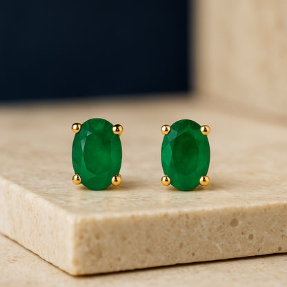 Pair of yellow metal stud earrings with oval green gemstones in prong settings, shown in a close-up front angle on a stone surface.