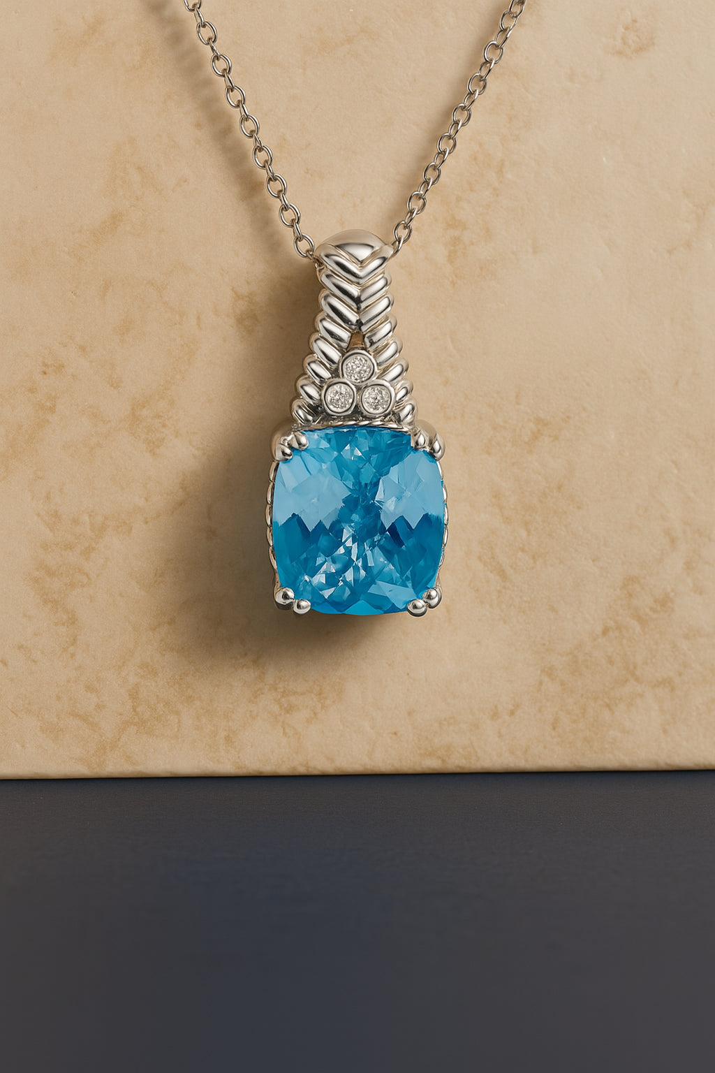 Close-up front view of a white necklace with a cushion-shaped blue gemstone in prong setting and three round white gemstones in bezel settings above it, displayed against a beige and dark blue background.