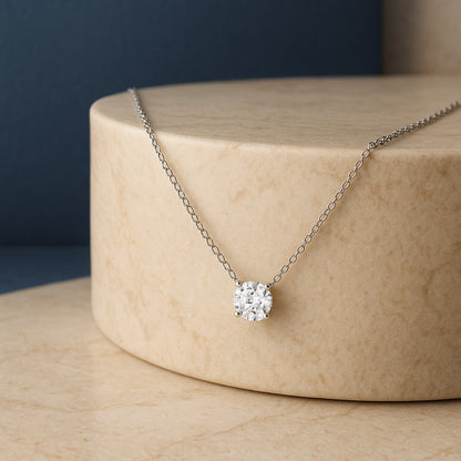 White metal necklace with a round white gemstone in a four-prong setting, displayed on a beige cylindrical pedestal, shown from the front angle.