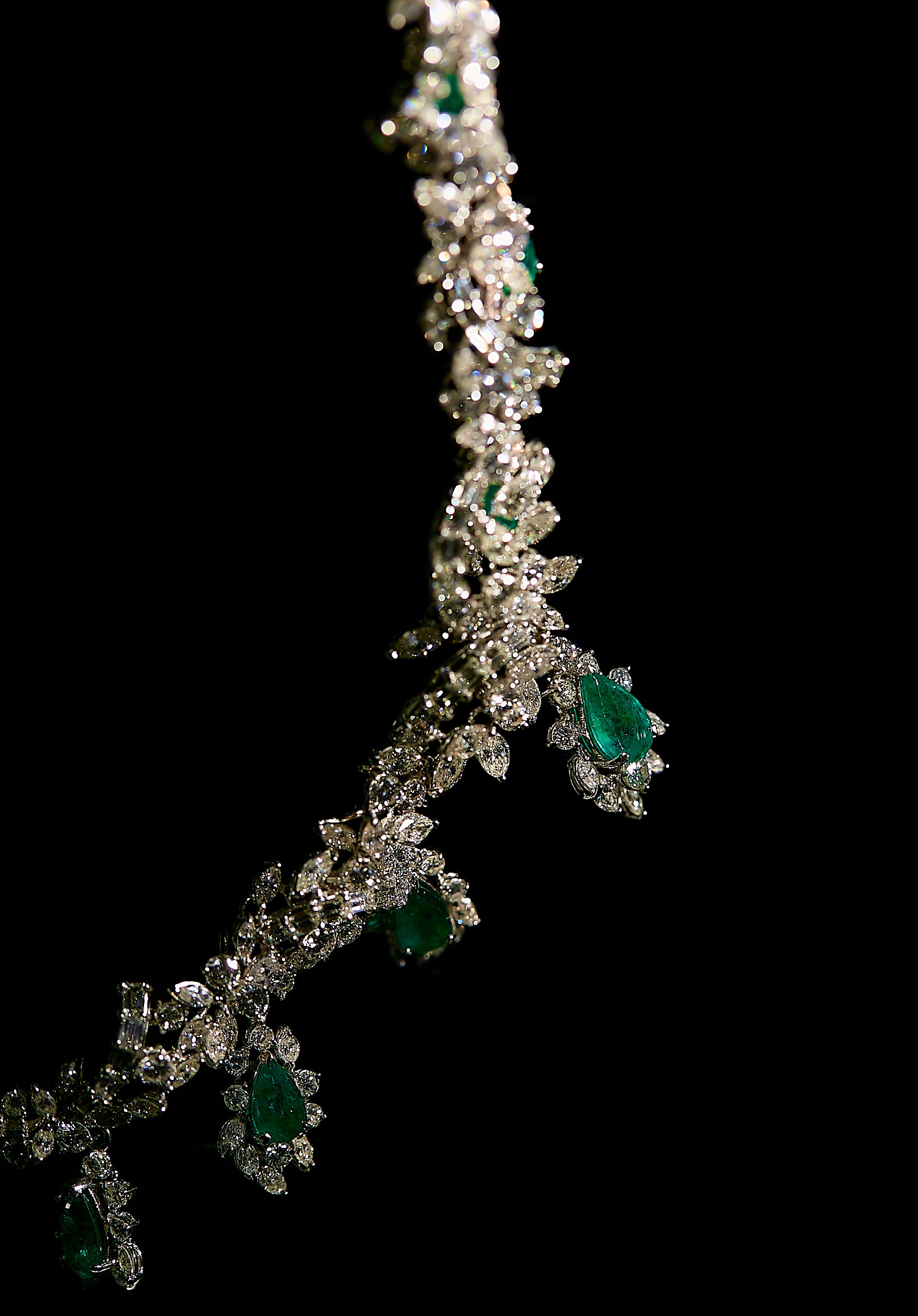 Close-up angled view of a white metal jewelry set featuring pear-shaped green gemstones surrounded by various shapes of white gemstones in prong settings, displayed against a black background.