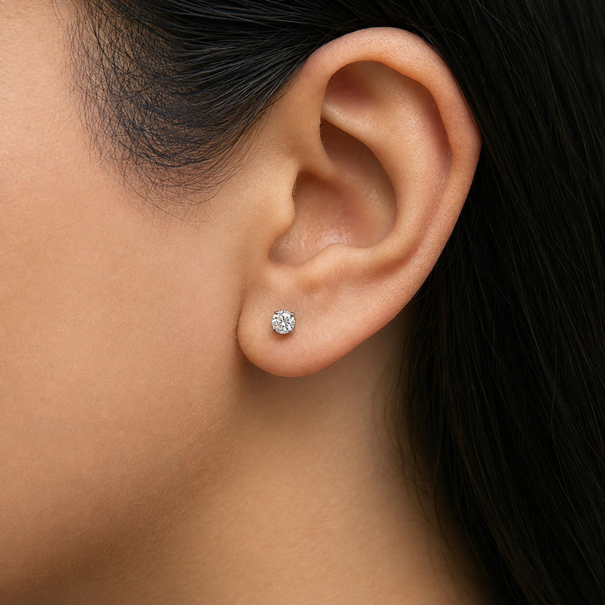 Close-up side view of a person's ear wearing a single round white gemstone stud earring in a four-prong white setting.