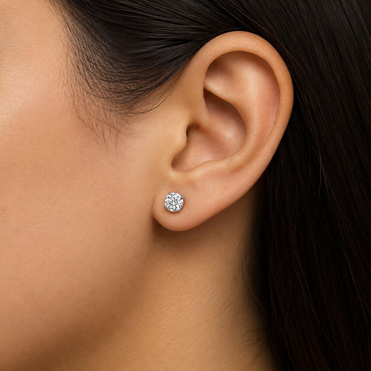 Close-up side view of a model's ear wearing a white metal earring with a round white gemstone set in a four-prong setting.