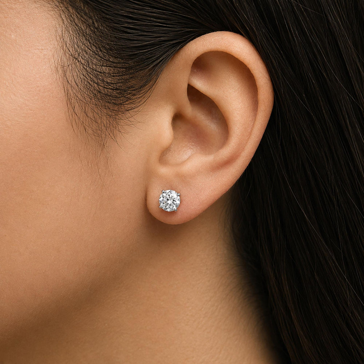 Close-up side view of a model's ear wearing white metal stud earrings with round white gemstones in a 4-prong setting.