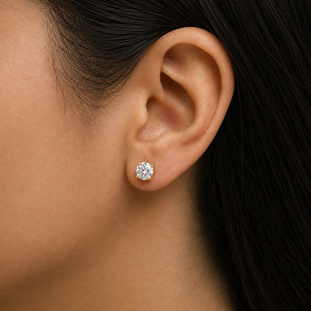 Close-up side view of a model wearing a single round white gemstone stud earring set in yellow metal with a 4-prong setting.