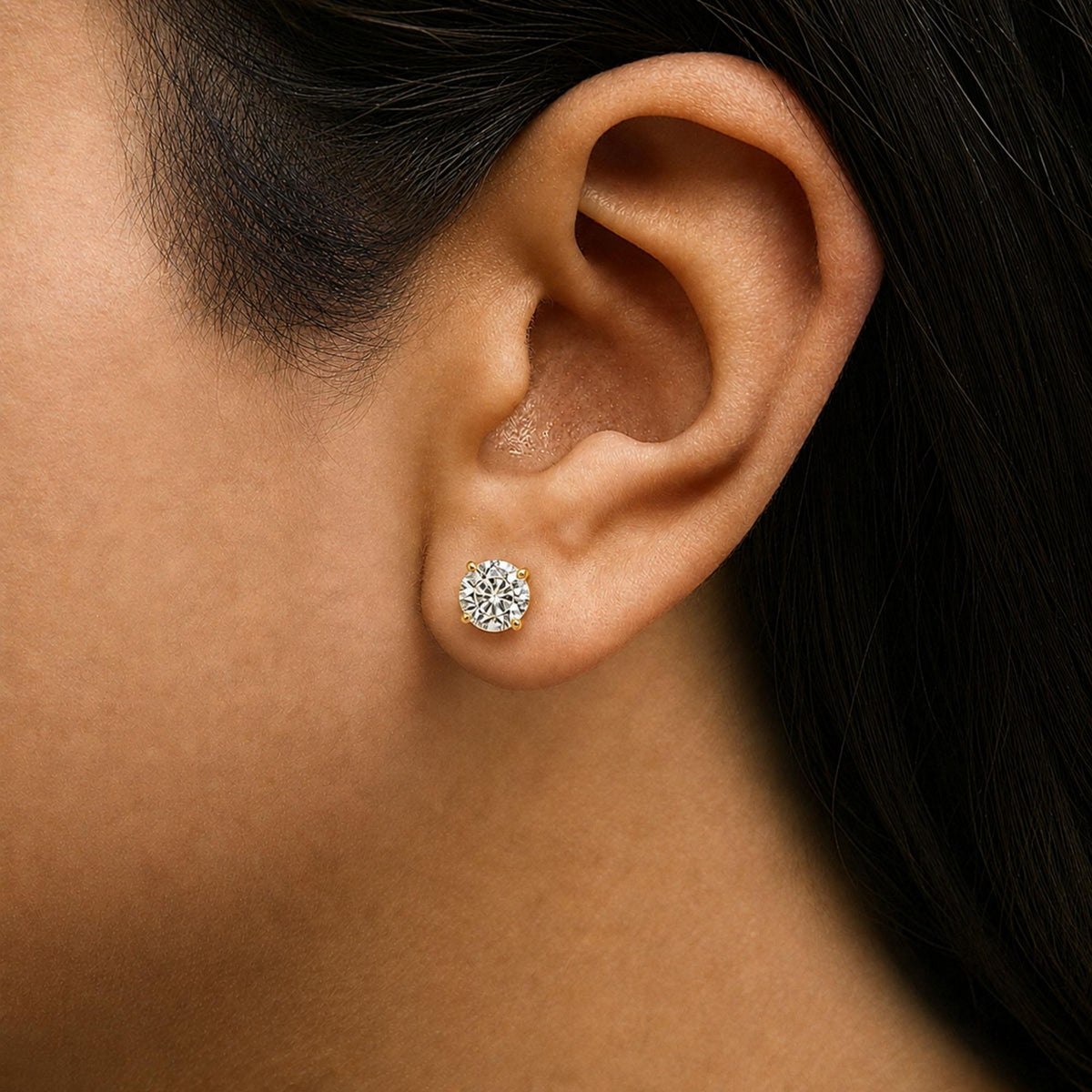 Close-up side view of a model wearing round white gemstone stud earrings set in yellow metal with four prongs.