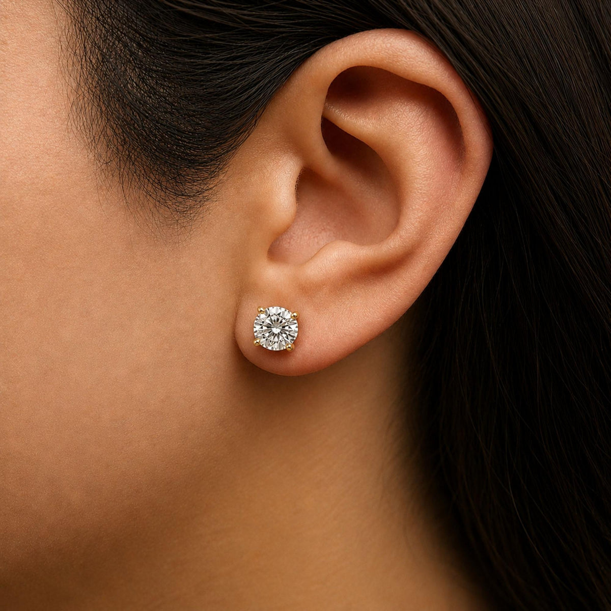 Close-up side view of a model wearing a yellow metal stud earring featuring a round white gemstone in a four-prong setting.