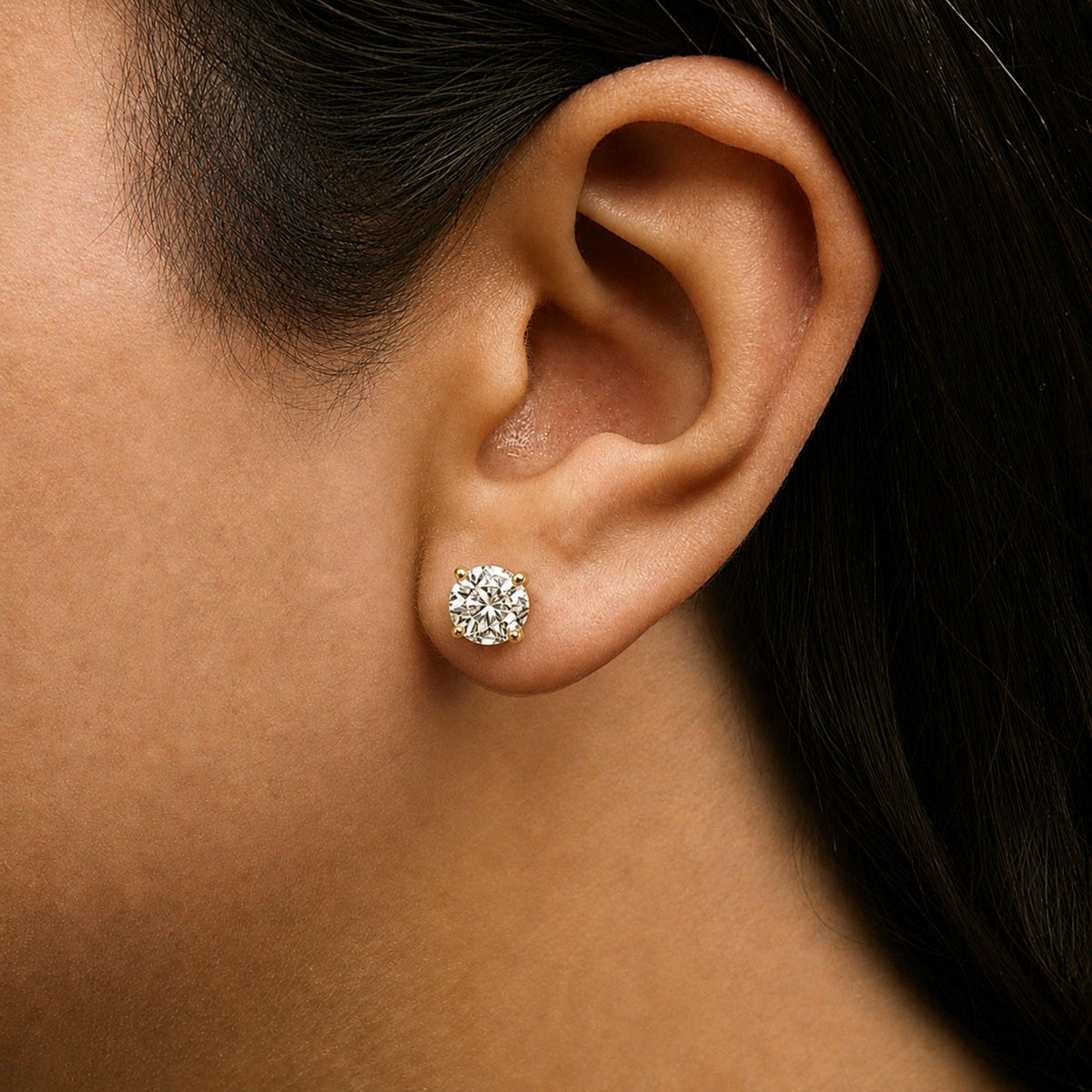 Close-up side view of a model wearing a pair of yellow metal stud earrings with round white gemstones in 4-prong settings.