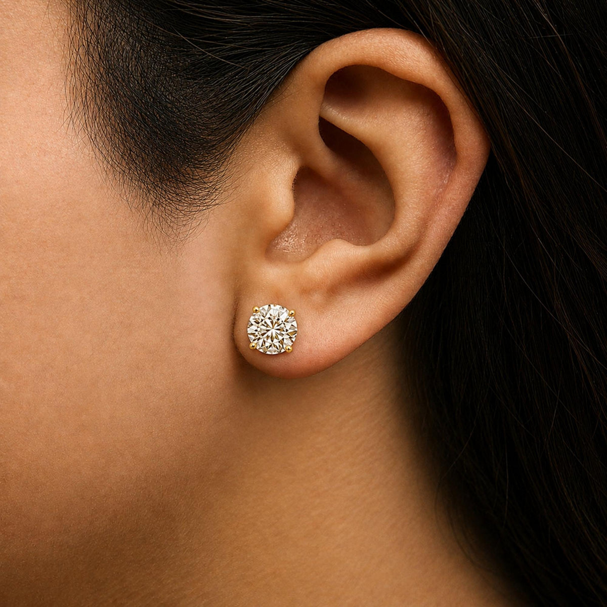 Close-up side view of a model wearing round white gemstone stud earrings set in yellow metal with four prongs.