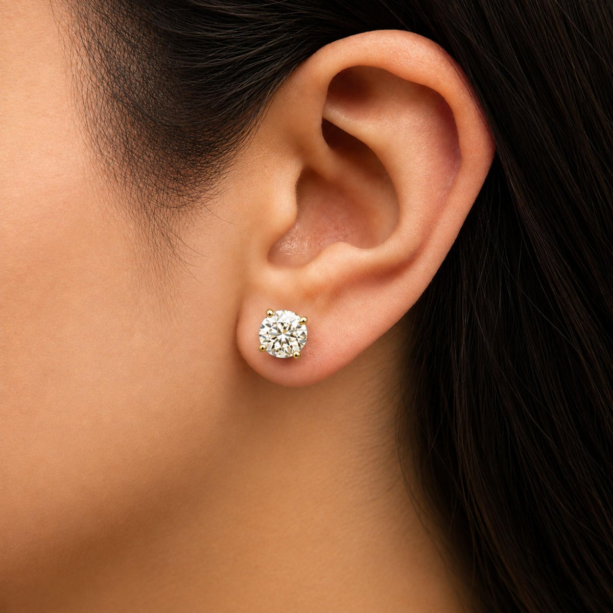 Close-up side view of a model wearing round white gemstone stud earrings set in yellow metal with 4-prong settings.