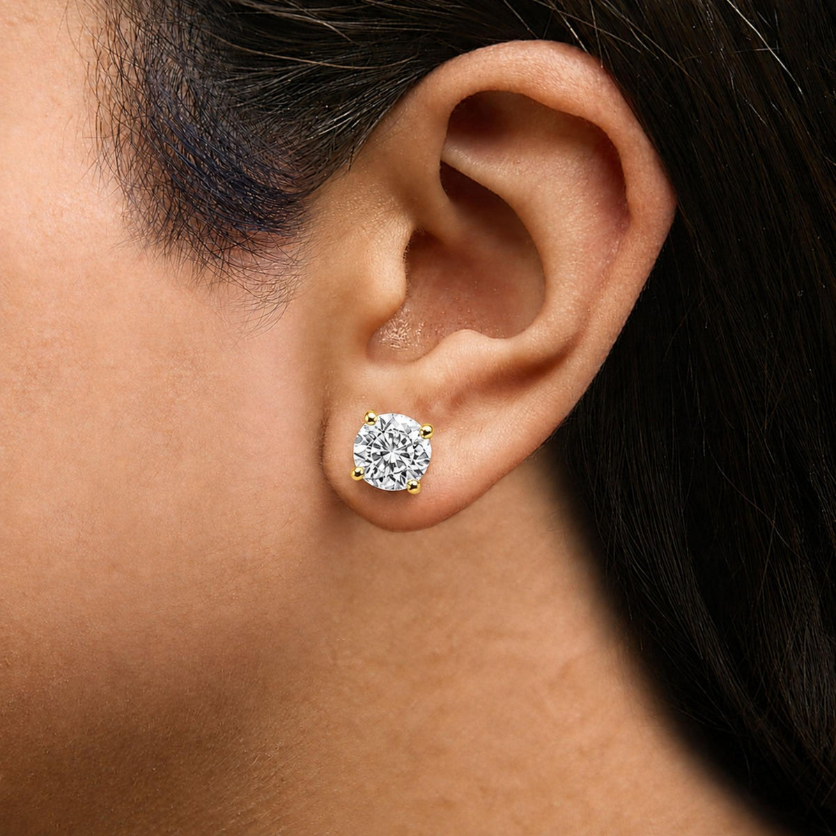 Close-up side view of a yellow metal earring with a round white gemstone in a four-prong setting, shown worn on a model's ear.