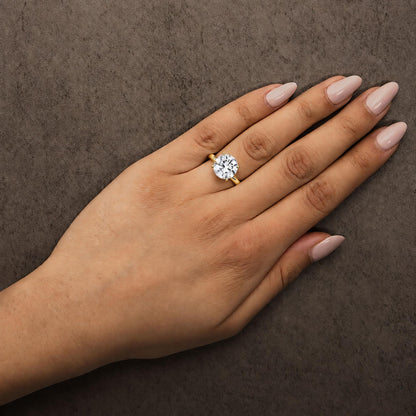 Yellow metal ring with a single round white gemstone in a 4-prong setting, shown worn on a model's finger from a top angle.