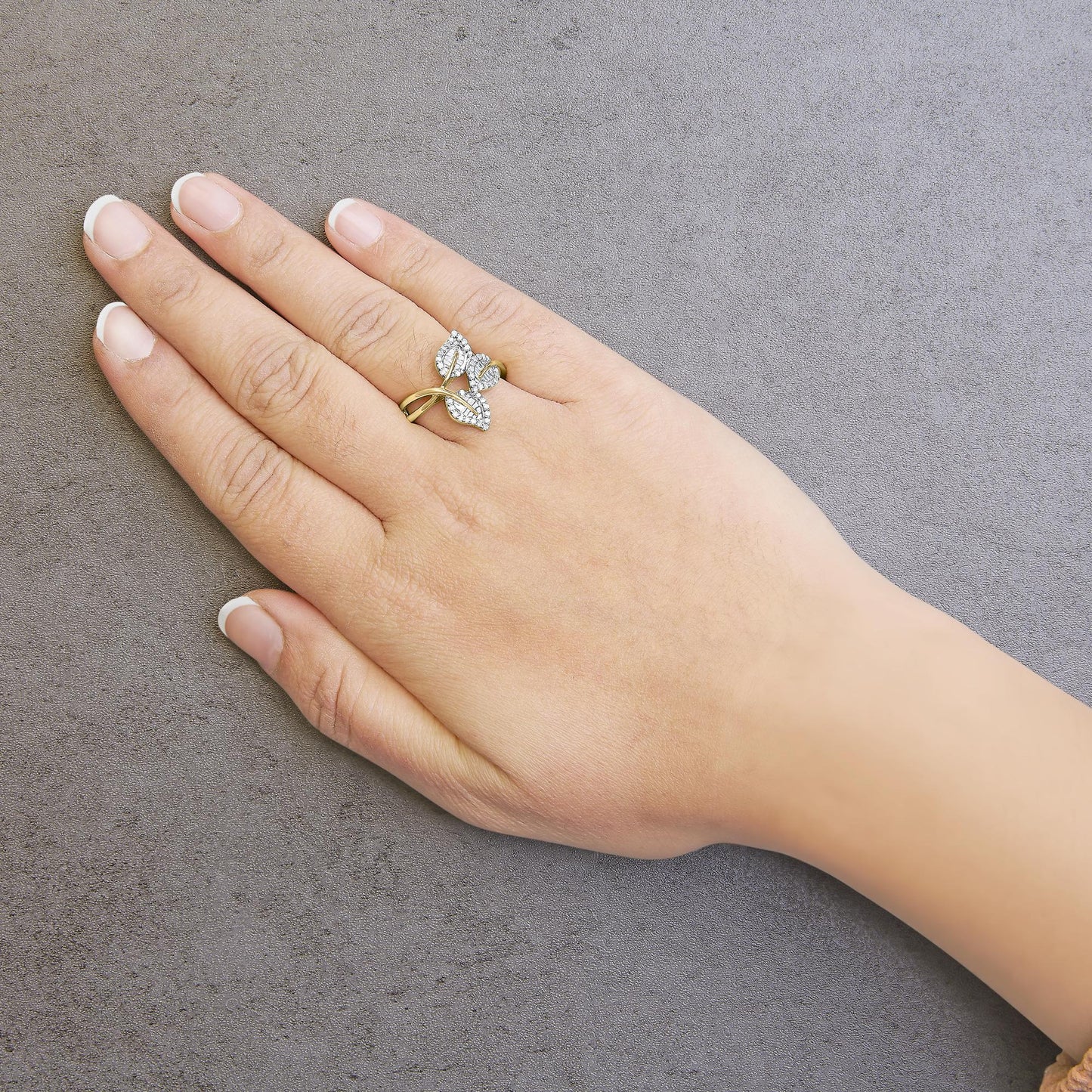 Close-up side angle of a yellow metal ring with three leaf-shaped sections featuring white round pave-set gemstones and white baguette channel-set gemstones, worn on a hand against a gray textured background.