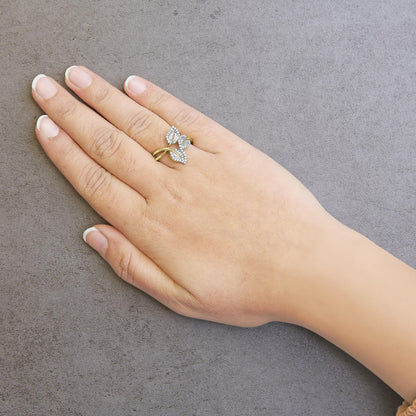 Close-up side angle of a yellow metal ring with three leaf-shaped sections featuring white round pave-set gemstones and white baguette channel-set gemstones, worn on a hand against a gray textured background.