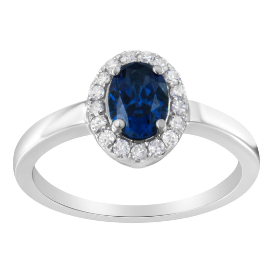 Front-facing shot of a white metal ring featuring an oval blue gemstone surrounded by a halo of round white gemstones in prong settings.