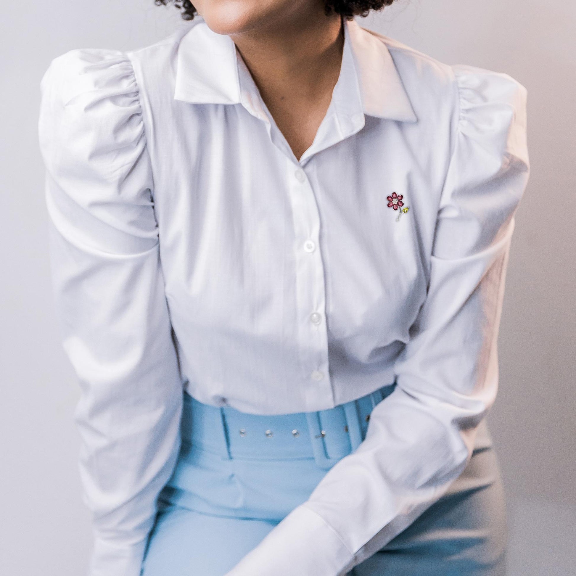 Close-up front angle of a white metal floral brooch with pink oval gemstones as petals, a green pear-shaped gemstone as a leaf, and white round gemstones as the stem, worn on a white shirt by a model.