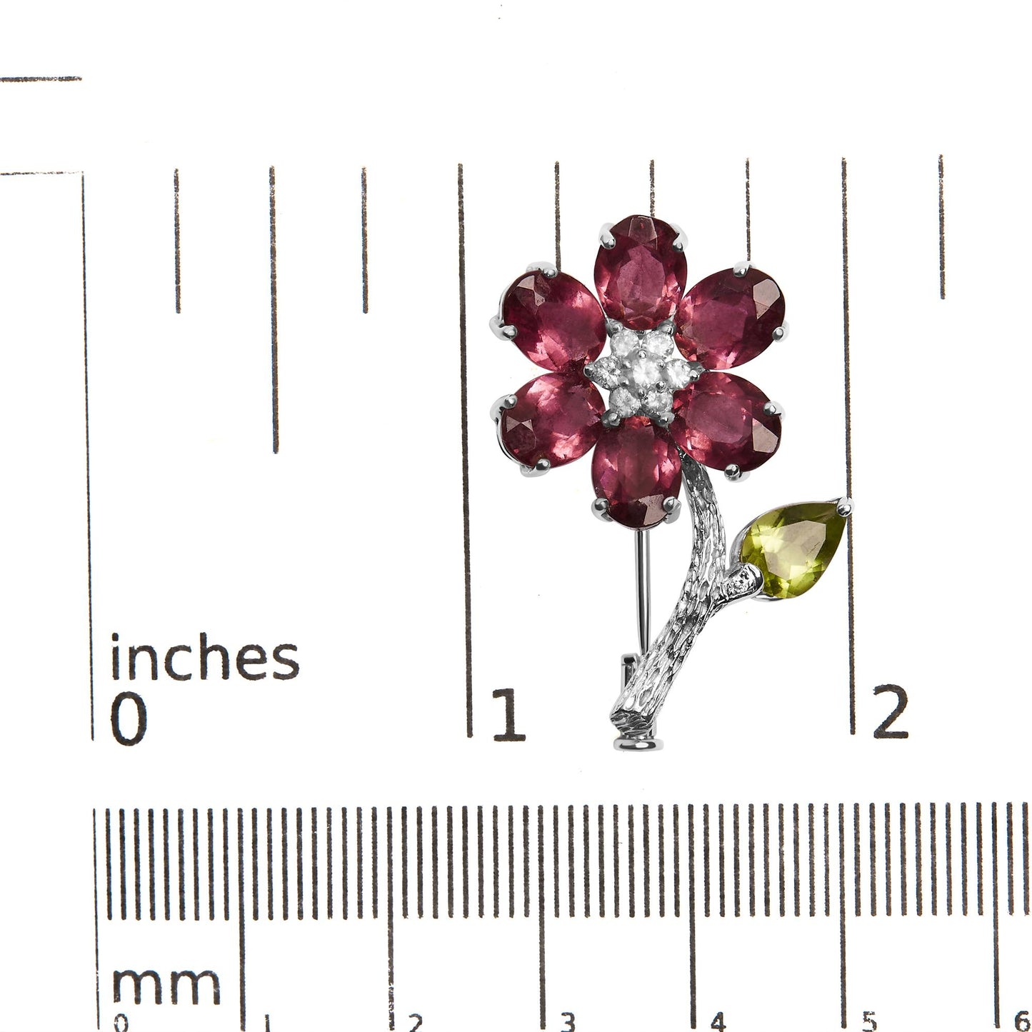Top-down view of a white metal flower-shaped brooch with six pink oval gemstones forming petals, a cluster of white round gemstones in the center, and a green pear-shaped gemstone as a leaf on the textured stem, placed on a ruler for size reference.