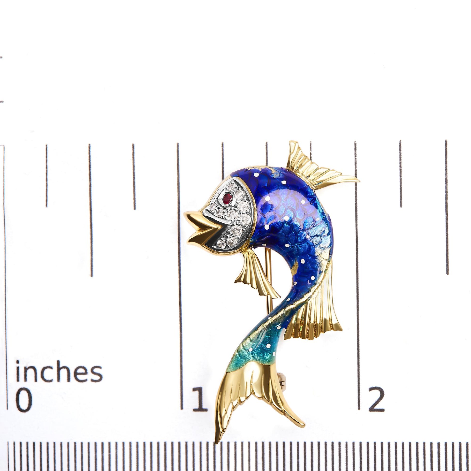 Yellow metal fish-shaped brooch with blue and turquoise enamel body, white round pave-set gemstones on the face, red round bezel-set gemstone eye, shown on a ruler for size reference, top-down angle.