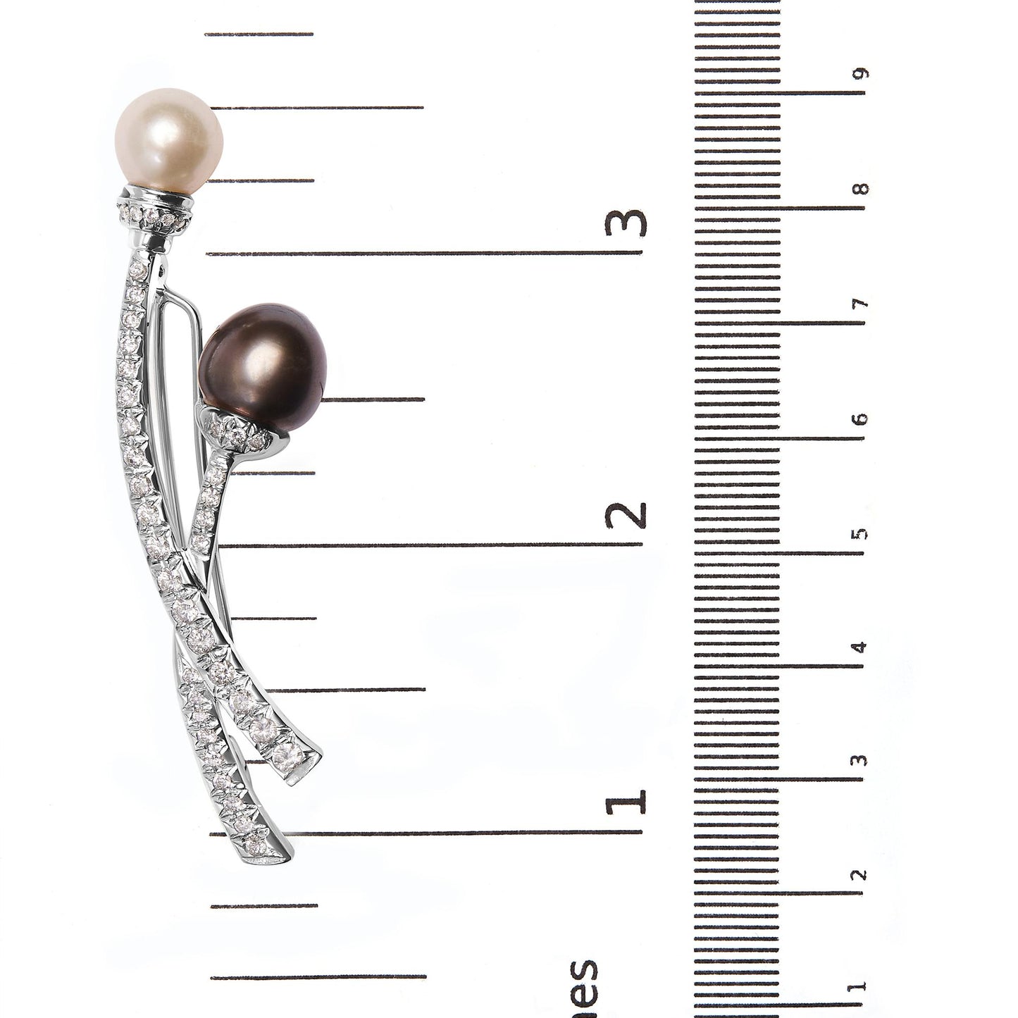 White metal brooch with two curved bands adorned with small white gemstones and topped with one white pearl and one dark pearl, positioned vertically on a ruler for size reference, front angle view.