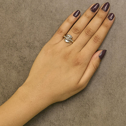 Yellow metal ring with one princess shape white gemstone in a prong setting, surrounded by baguette and round white gemstones in channel settings, shown worn on a model's hand from a top-down angle.