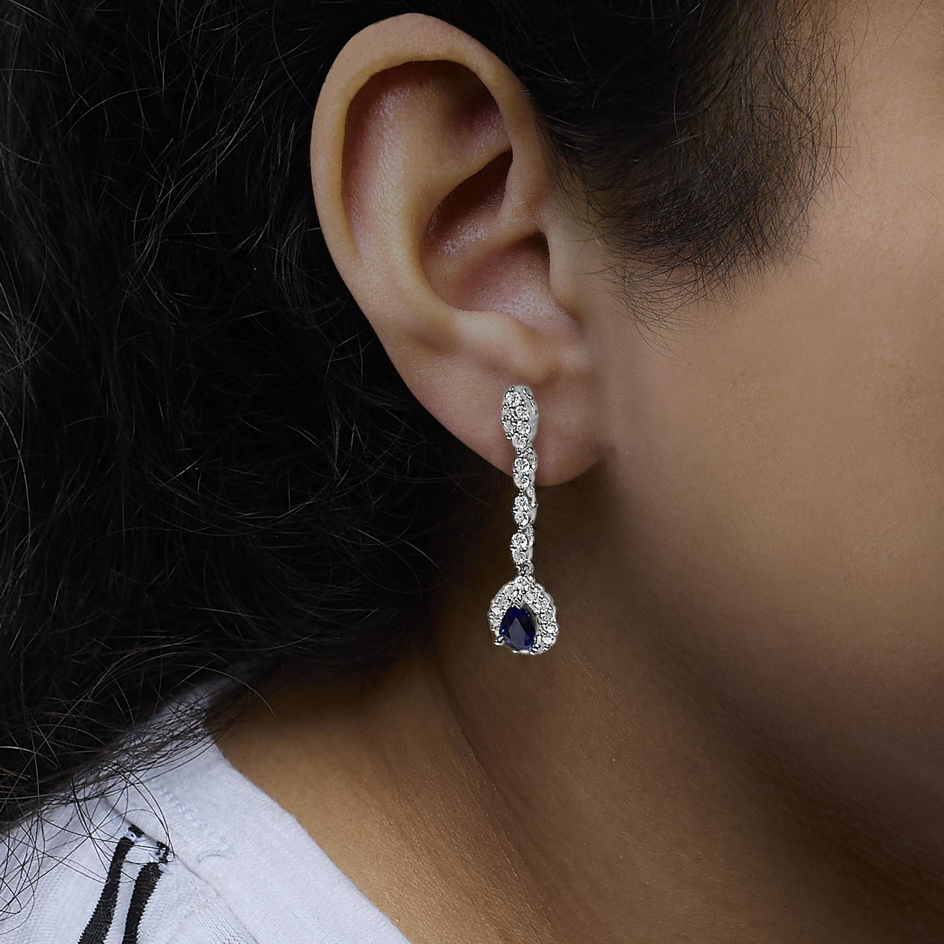 Close-up side view of a white metal dangling earring on a model's ear, featuring multiple round white gemstones in prong settings leading to a pear-shaped blue gemstone surrounded by smaller white gemstones.