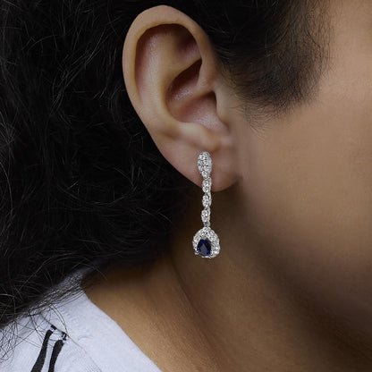 Close-up side view of a white metal dangling earring on a model's ear, featuring multiple round white gemstones in prong settings leading to a pear-shaped blue gemstone surrounded by smaller white gemstones.