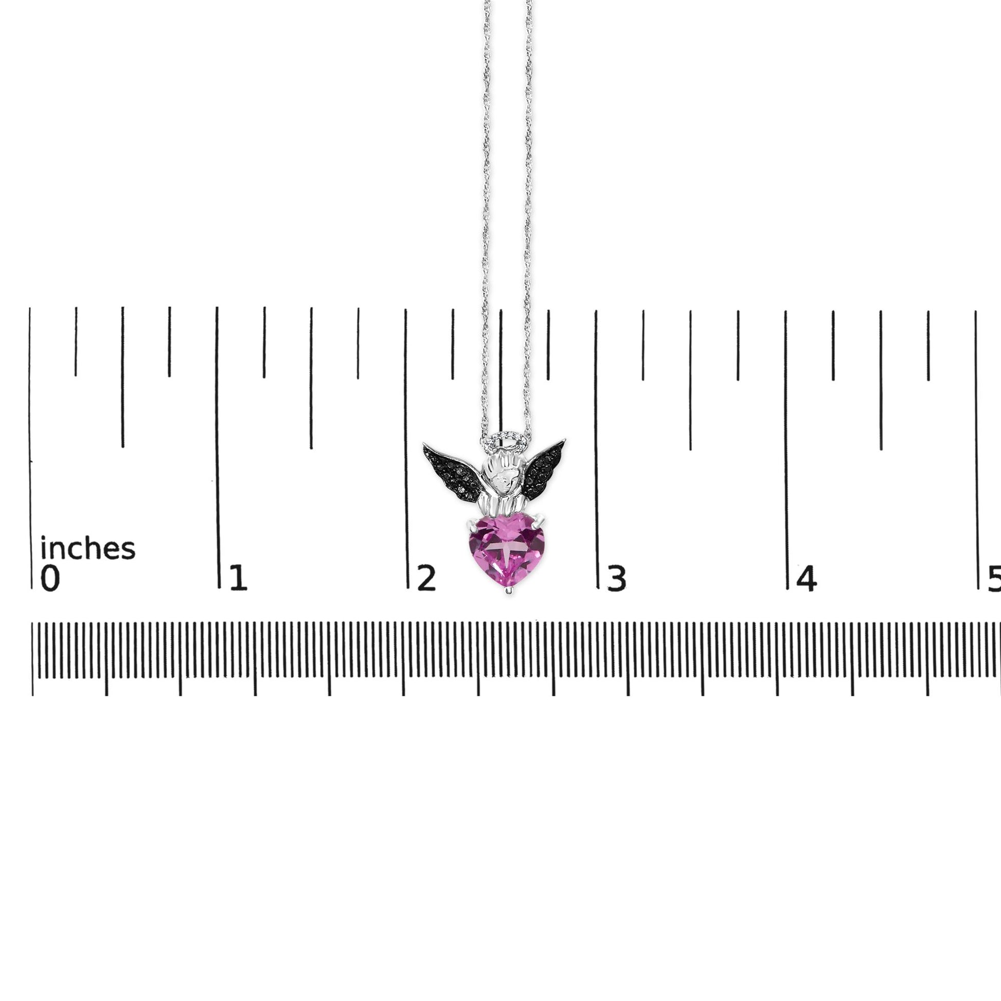 Necklace shown on a scale, photographed from the front, featuring a white and black metal angel pendant with a heart-shaped pink gemstone and round white and black gemstones.